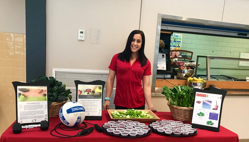UMass Lowell alumni Michelle Palladino pictured in a school cafeteria with a table full of healthy snacks and information on leading a healthy lifestyle