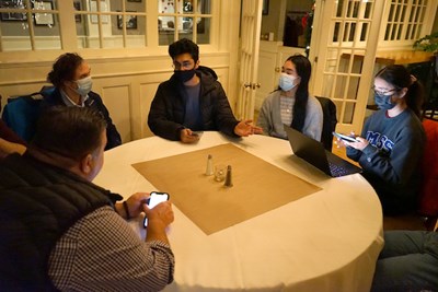 Three students and a professor wearing masks sit around a restaurant table with a man holding a phone