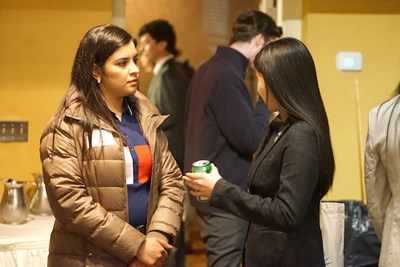 Two young women talk to each other in a room with people milling around in the background