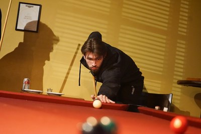 A young man plays pool in a room with the shadow of a person on the wall behind him