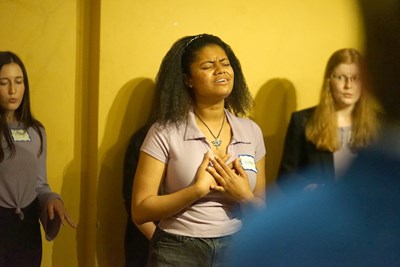 A young woman closes her eyes while singing in a room as two other women look on