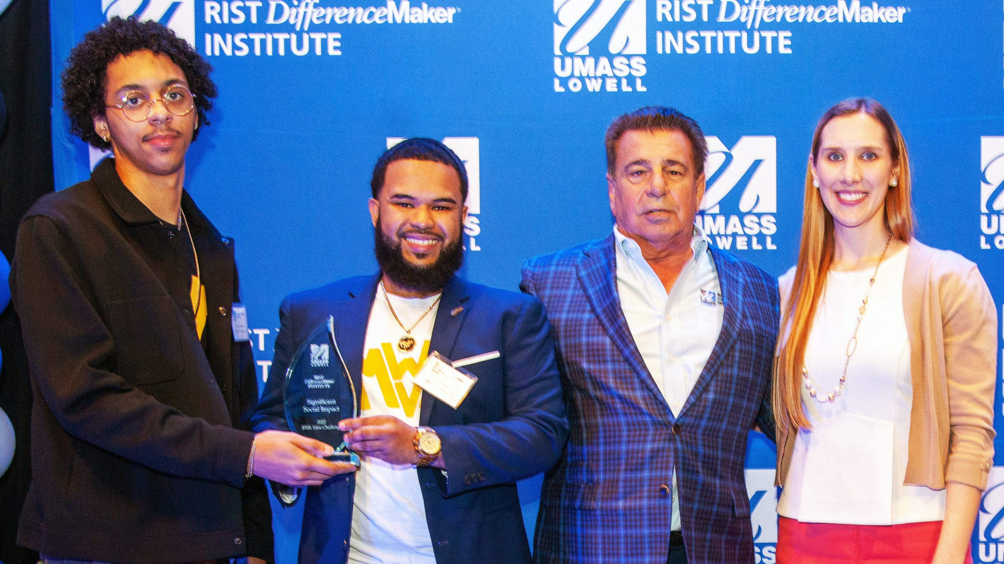 2 male students from the Minds With Purpose team holding an award pose with Brian Rist and Holly Lalos of Difference Makers against a blue UMass Lowell backdrop.