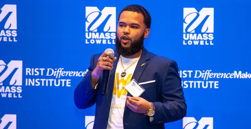 One of the members Minds With Purpose team holding a microphone and speaking in front of blue UMass Lowell backdrop.