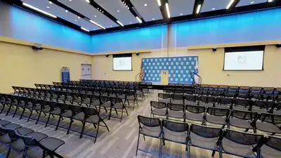 Wide view of a large room with arrays of foldable chairs, all facing a podium on a well-lit stage.