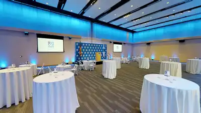 Large room with ten tall tables and five short tables, each draped in a white tablecloth; the short tables are each surrounded by four chairs.