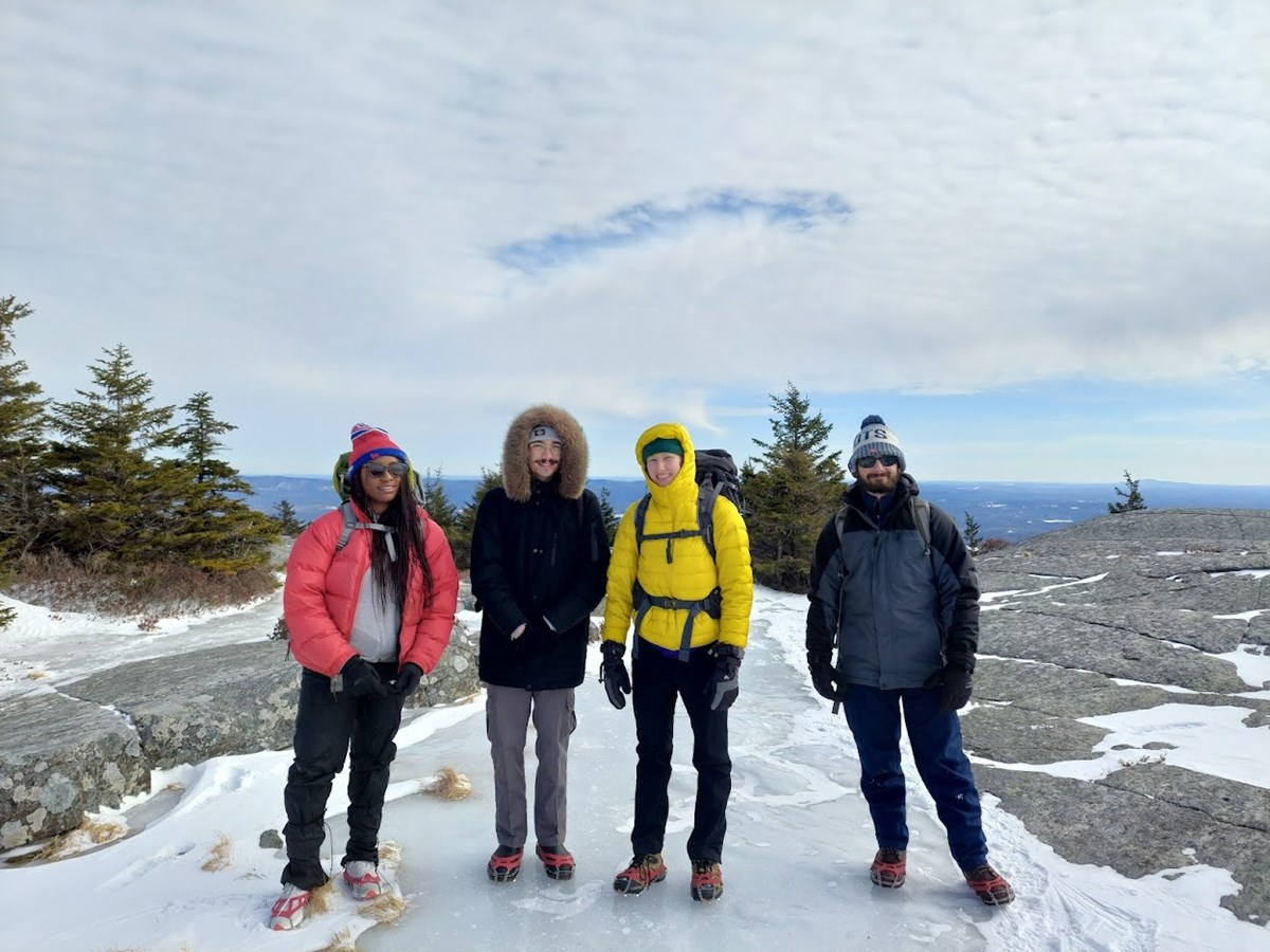four hikers smile while standing on an snowy patch near a mountain summit