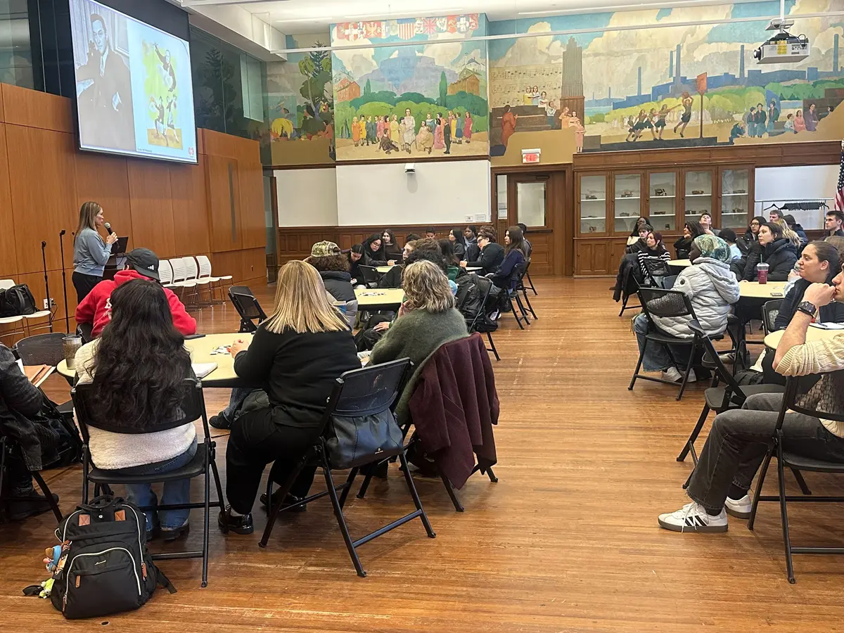 Students sit at tables in large hall for workshop presentation.