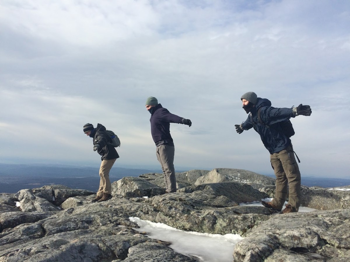 Three male-presenting students with arms spread like they were flying on top of the windy summit of Mt. Monadnock.