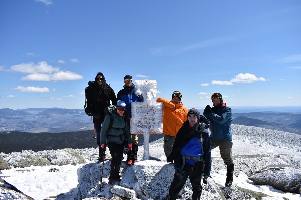 A group of happy hikers stands atop the summit of Mt. Moosilauke near a iced over marker sign with a dramatic snow covered mountain side in the background