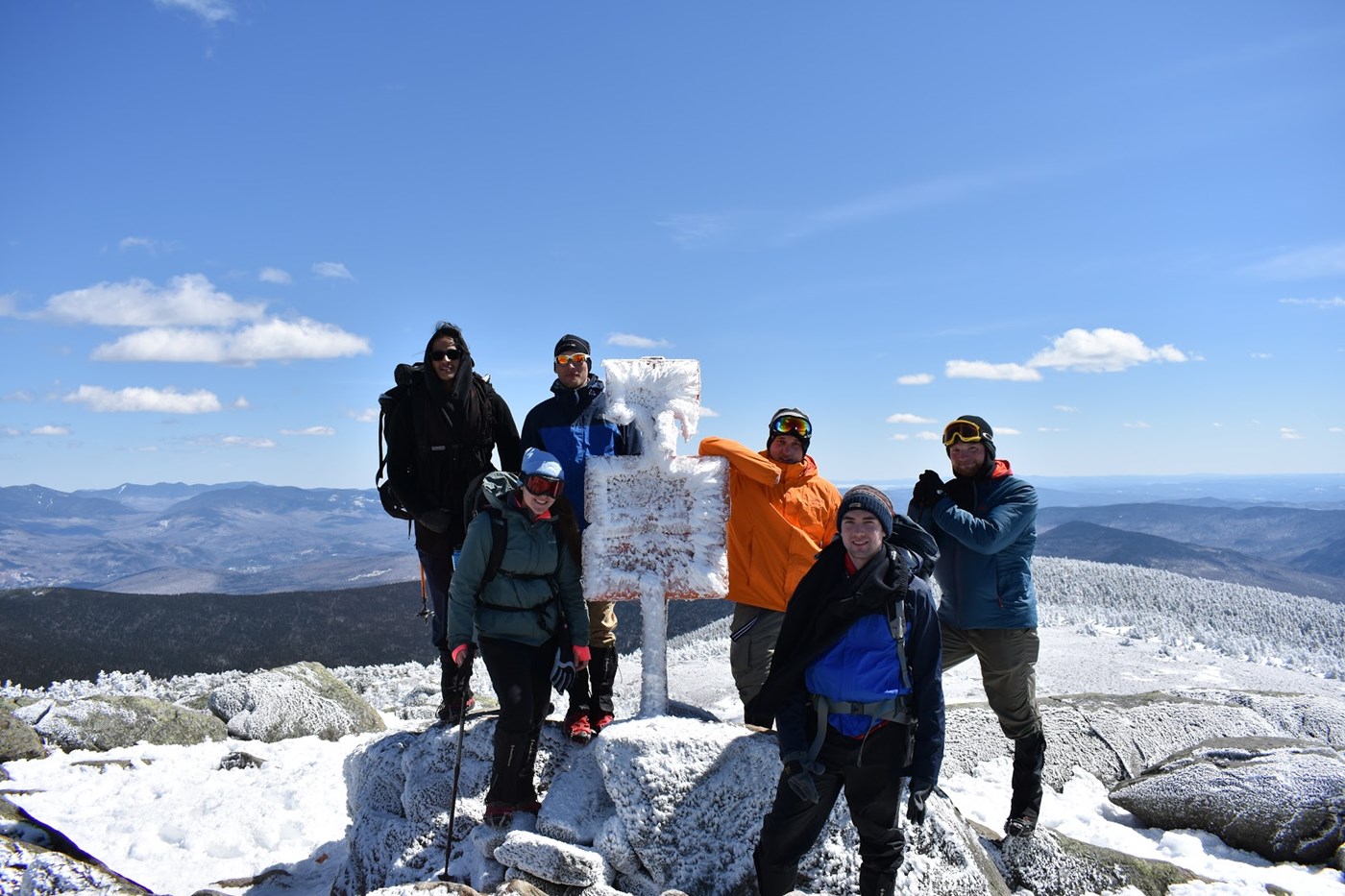 Group of hikers standing atop Mount Moosilauke’s summit near an iced over marker sign with a snow covered mountain side in the background.