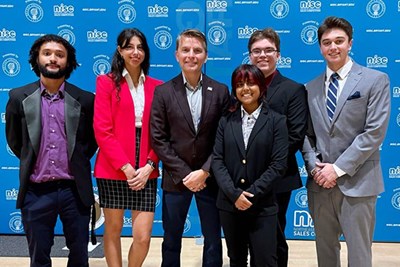 Five students and a professor in business attire pose for a group photo in front of a blue backdrop