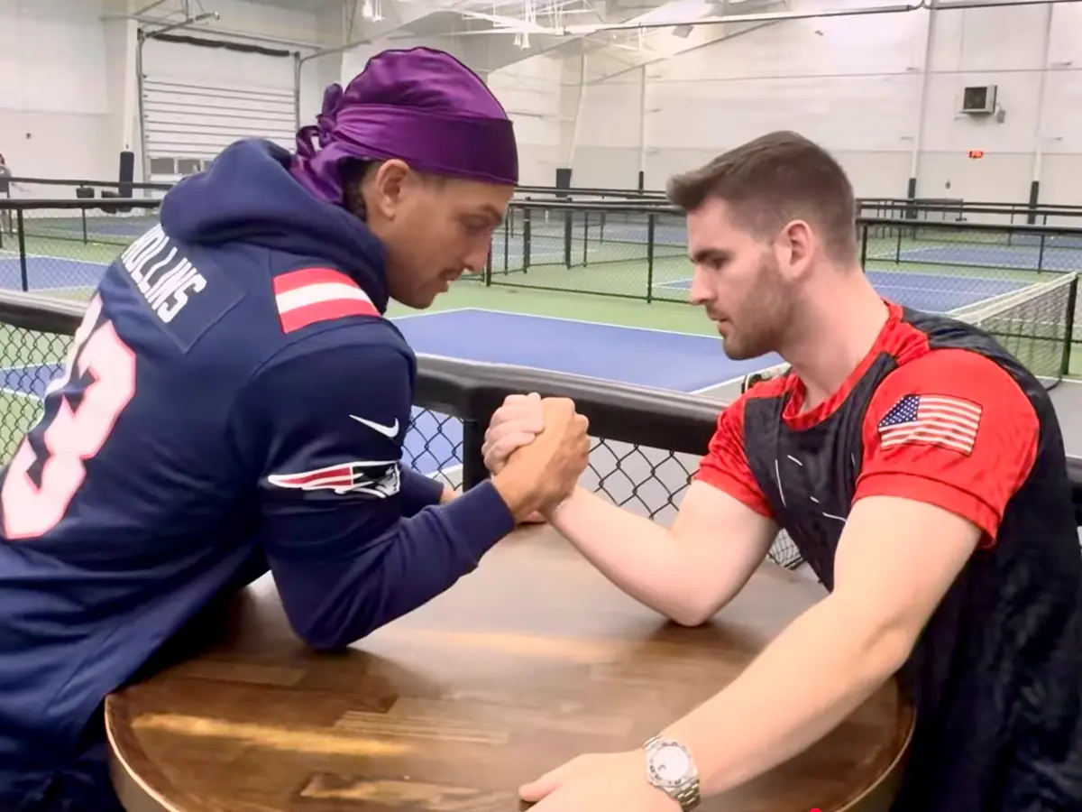A man in a football jersey and purple head covering arm wrestles a man inside a pickleball facility.