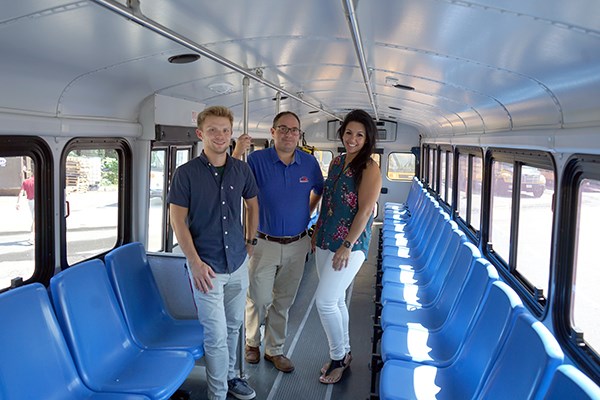 Two men and a woman stand in the middle of a bus with blue seats
