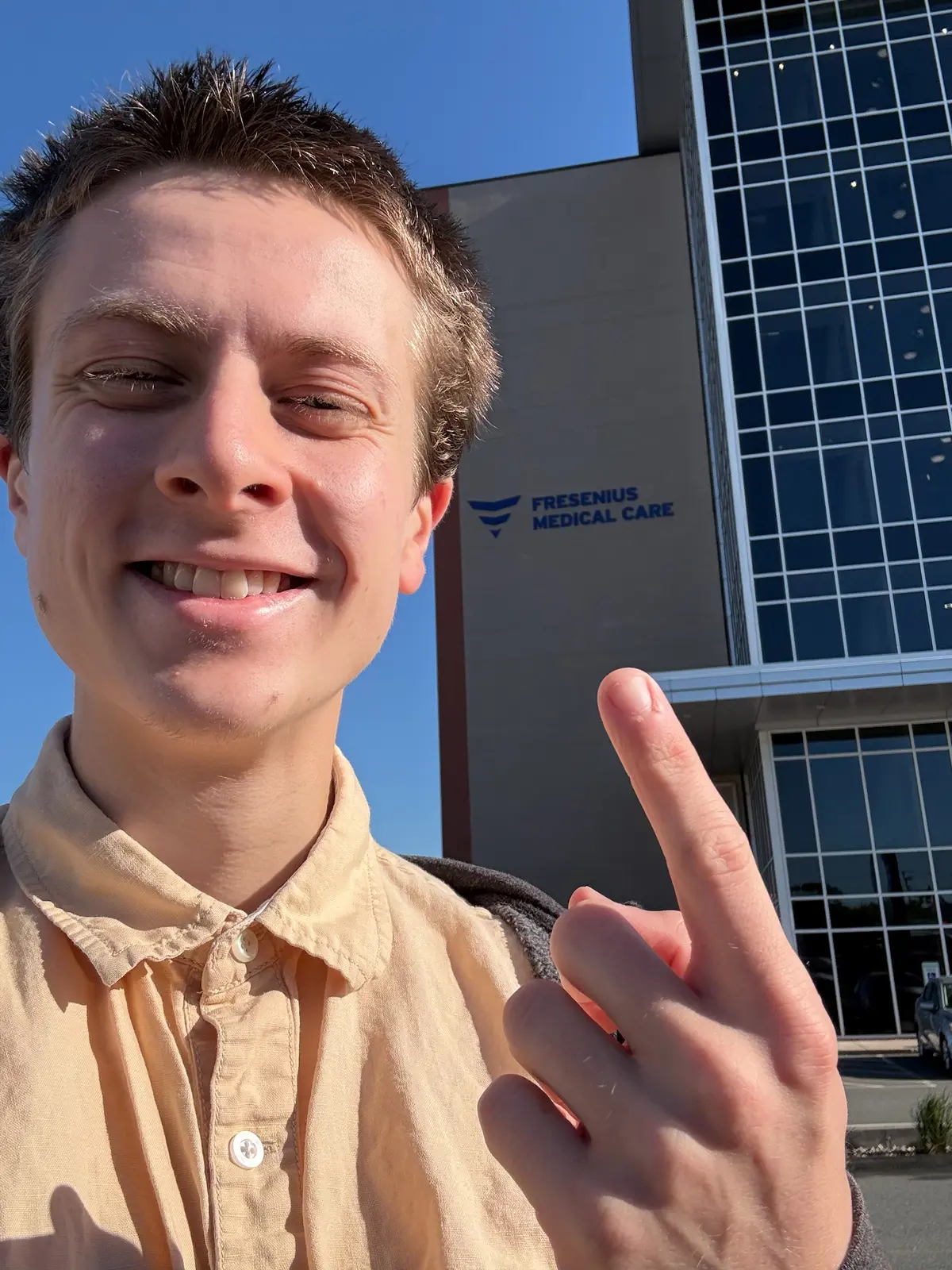 A young man points to the front of a building while taking a selfie outside.