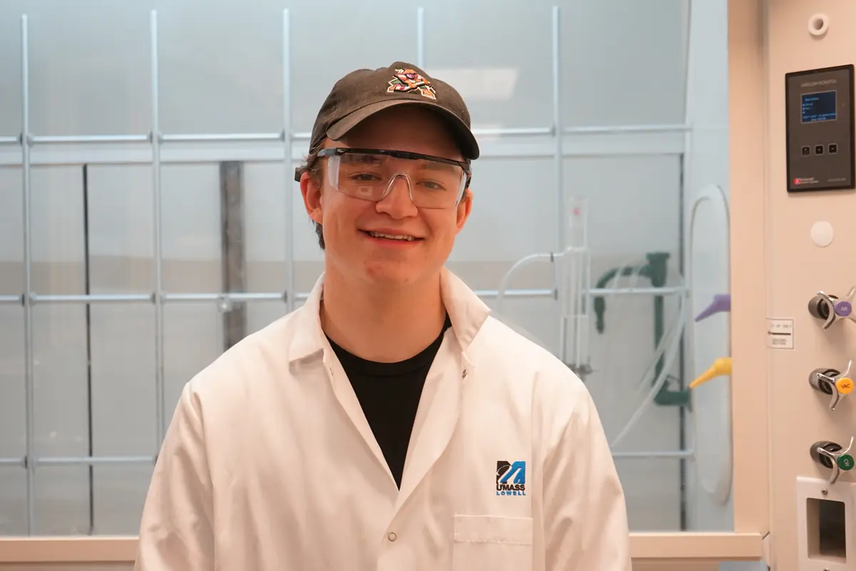 A young man wearing a lab coat, safety glasses and baseball cap poses for a photo.