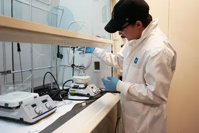 A young man in a white lab coat and dark cap works at a chemistry bench.