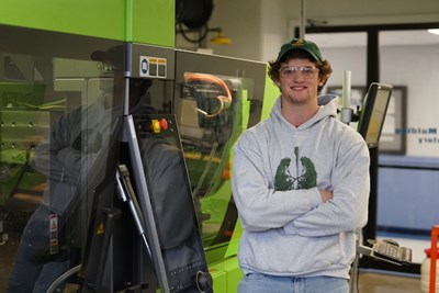 A young man in a hat and safety goggles poses for a photo next to a plastics machine
