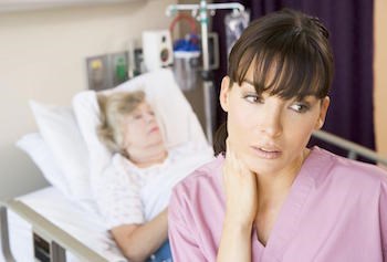 Image of a nurse in foreground looking stressed with an older woman patient lying in a hospital bed behind her. The image depicts the need for the job stress program which would help nurses learn how to minimize stress, which would in turn translate to improved patient care.