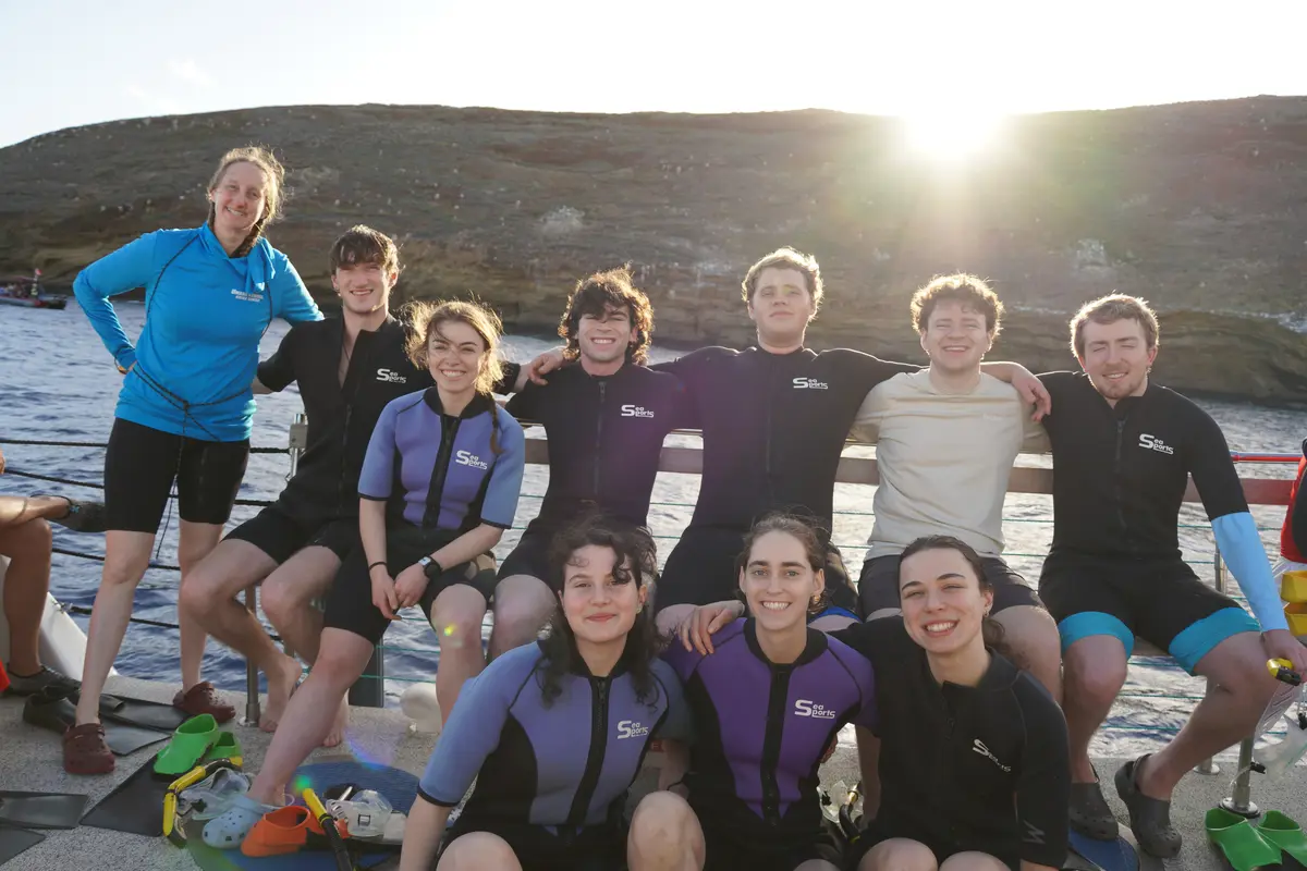Ten people pose for a group photo while sitting on a boat with the sun setting behind them.