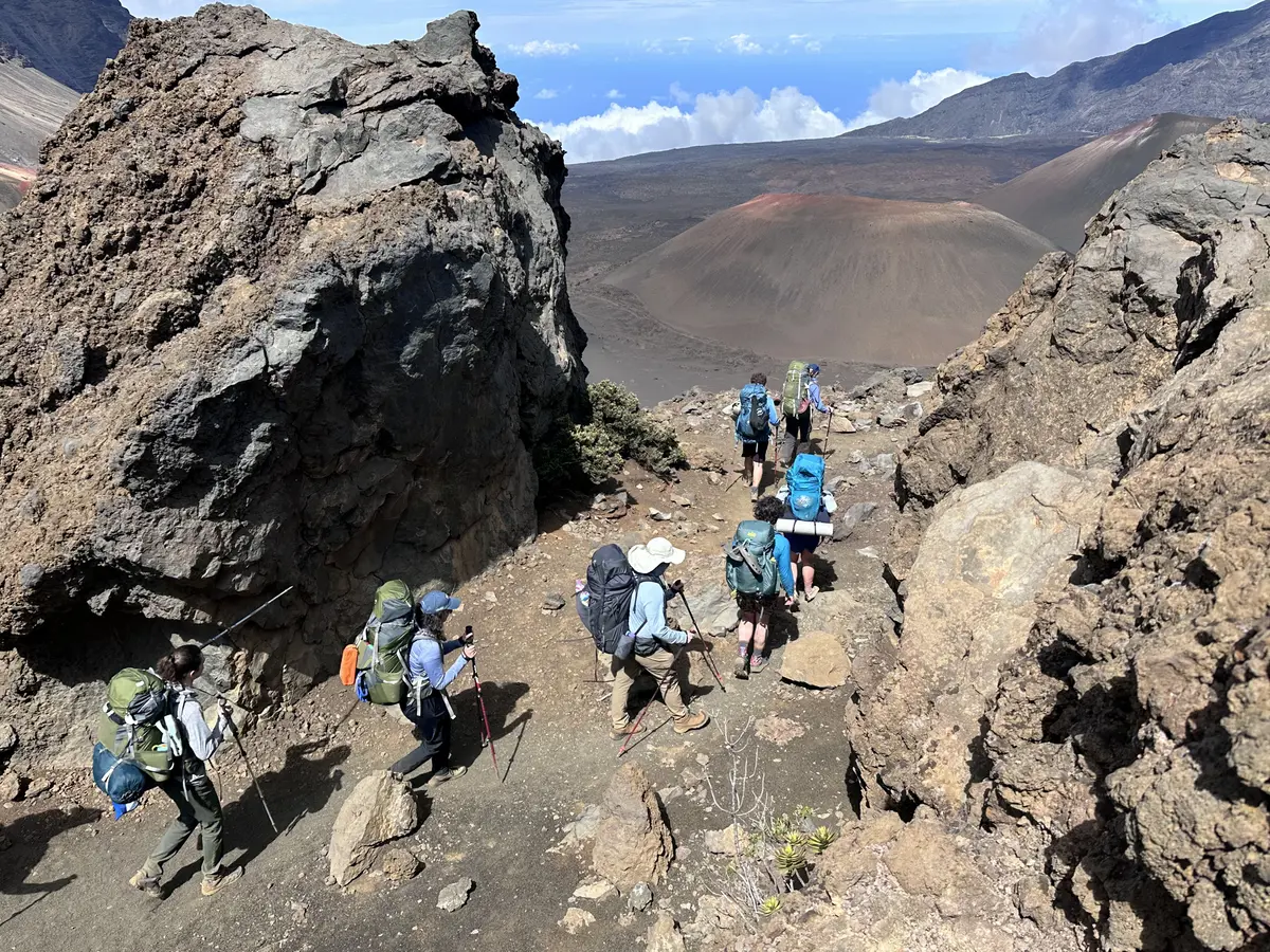 Seven people hike across a rocky terrain with a volcano crater in front of them.