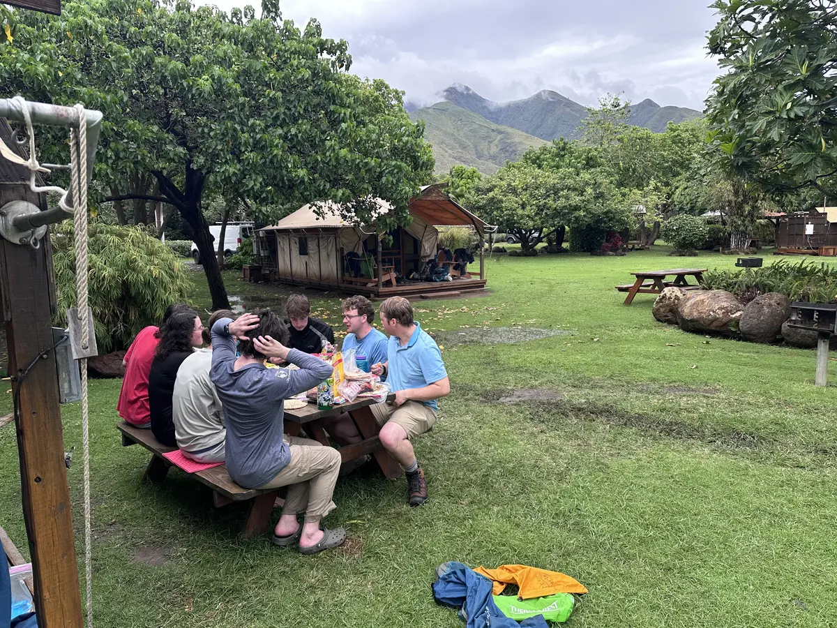 Seven people sit at a picnic table at a camp site in a lush tropical setting.