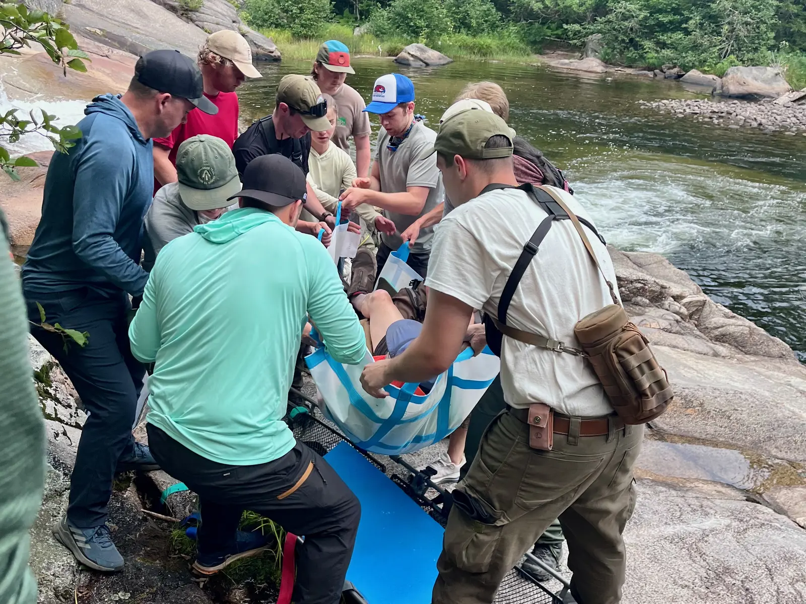 Nine people work together to load a man into a litter alongside a river in the woods.