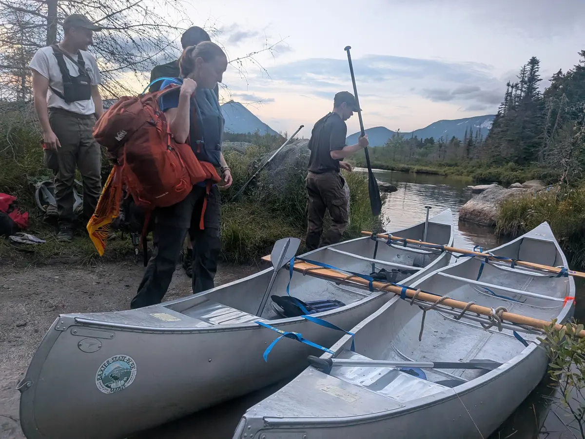 Four people approach two canoes that are tied together alongside a river in the wilderness.