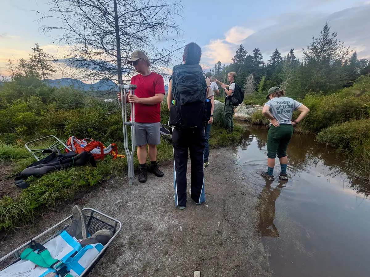 A half dozen people stand around while helping to evacuate a man in a litter in the outdoors.