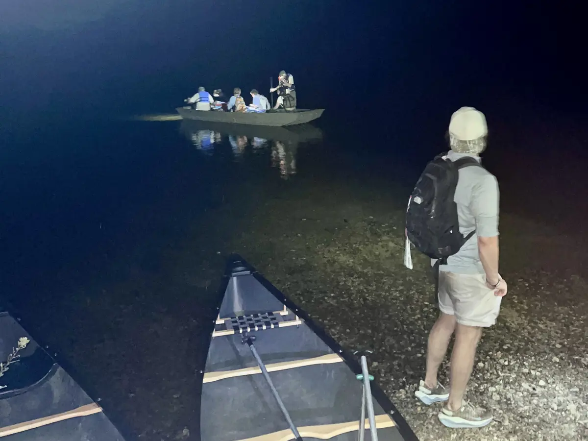 A man looks on from the shore while a boat with several people in it approaches on a pond at night.