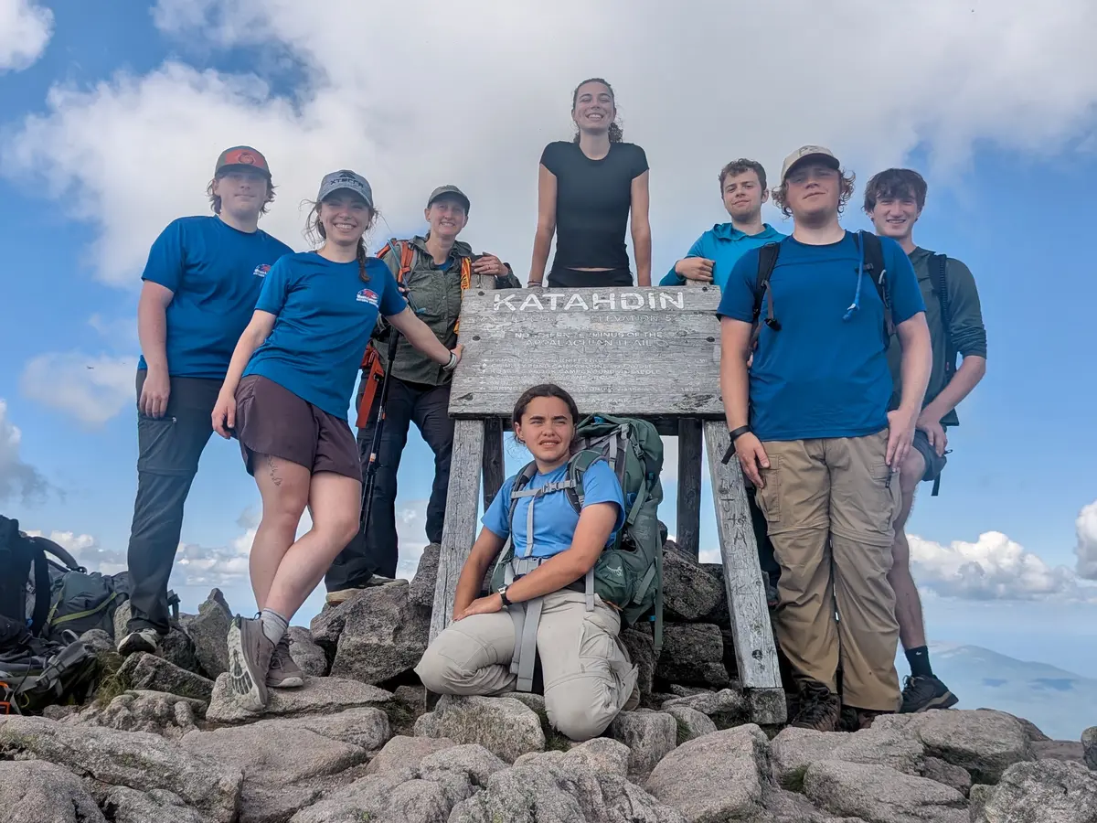 Eight men and women pose for a group photo next to a sign at the top of a mountain.