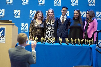 A man takes a photo of five people who are standing behind a table with dozens of small trophies on it