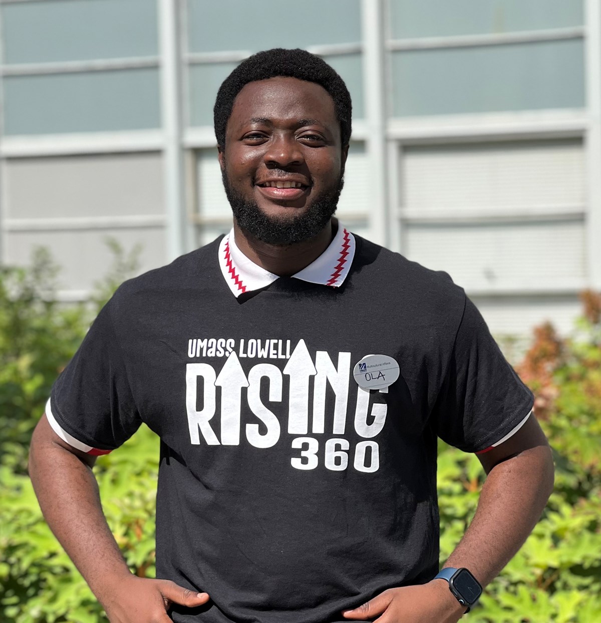 Olasunkanmi Olorunsaye standing outside wearing a shirt with text: UMass Lowell Rising 360 and a name tag with OLA on it.