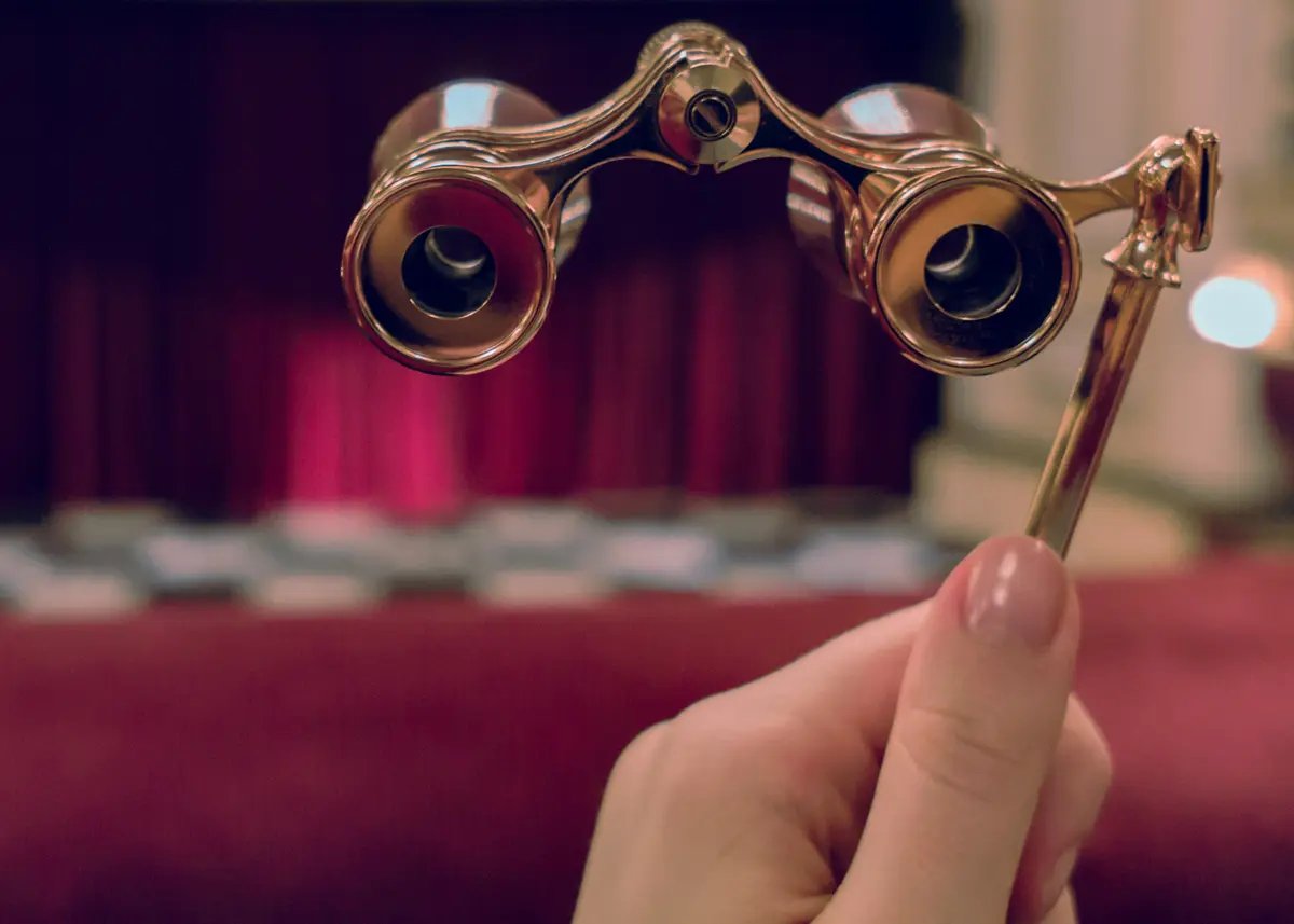 Hand holds opera glasses in theater against stage and drop-curtain.