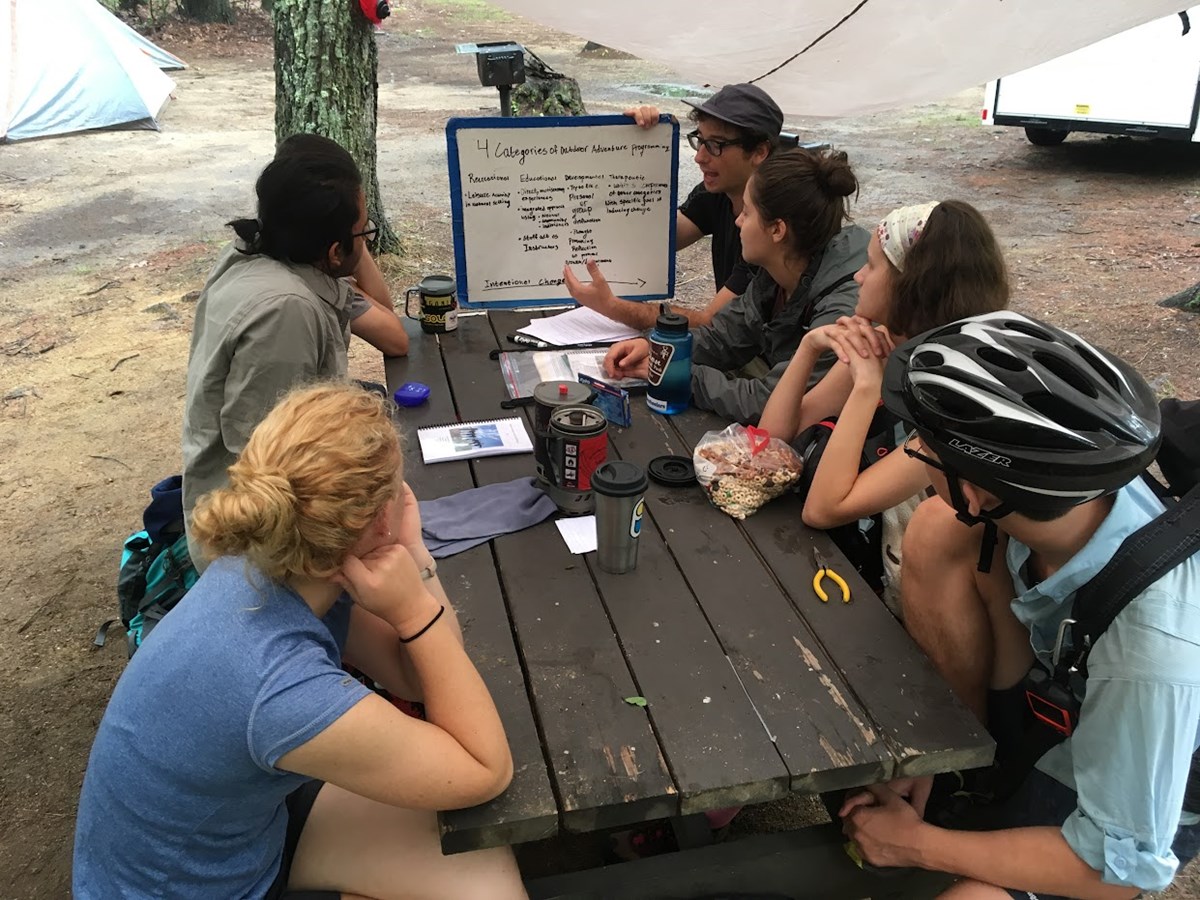 a group sits outside at a picnic table looking at a white board