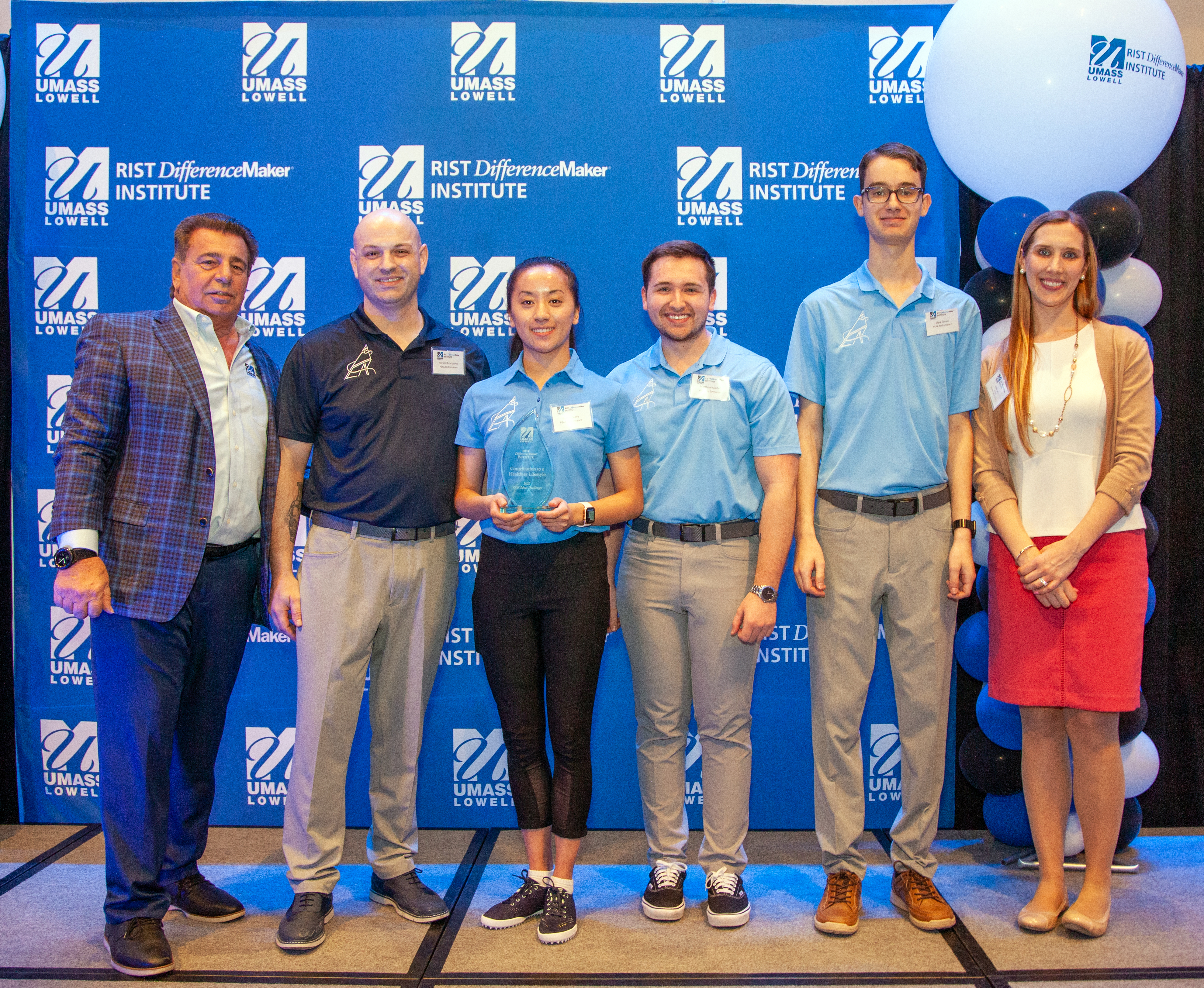3 male students and 1 female from the PEAK Performance team holding an award pose with Brian Rist and Holly Lalos of Difference Makers against a blue UMass Lowell backdrop.