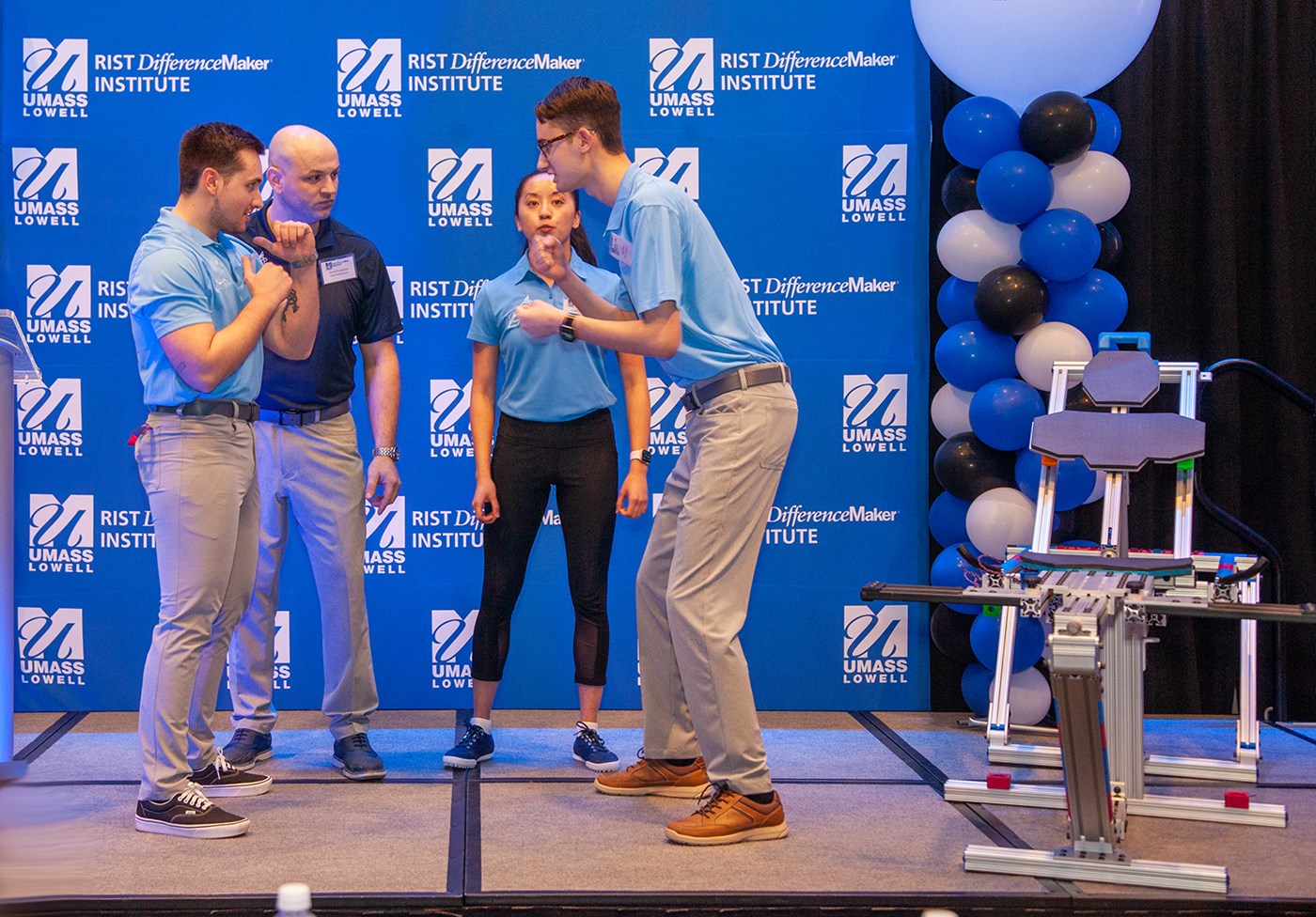 The PEAK Performance team standing in a group of 4 looking at each other in front of a blue UMass Lowell backdrop with their device off to the side.