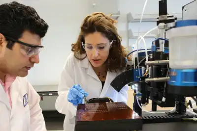 A woman in a lab coat and safety glasses uses tweezers on electronics in a lab while a man in a lab coat and glasses looks on.