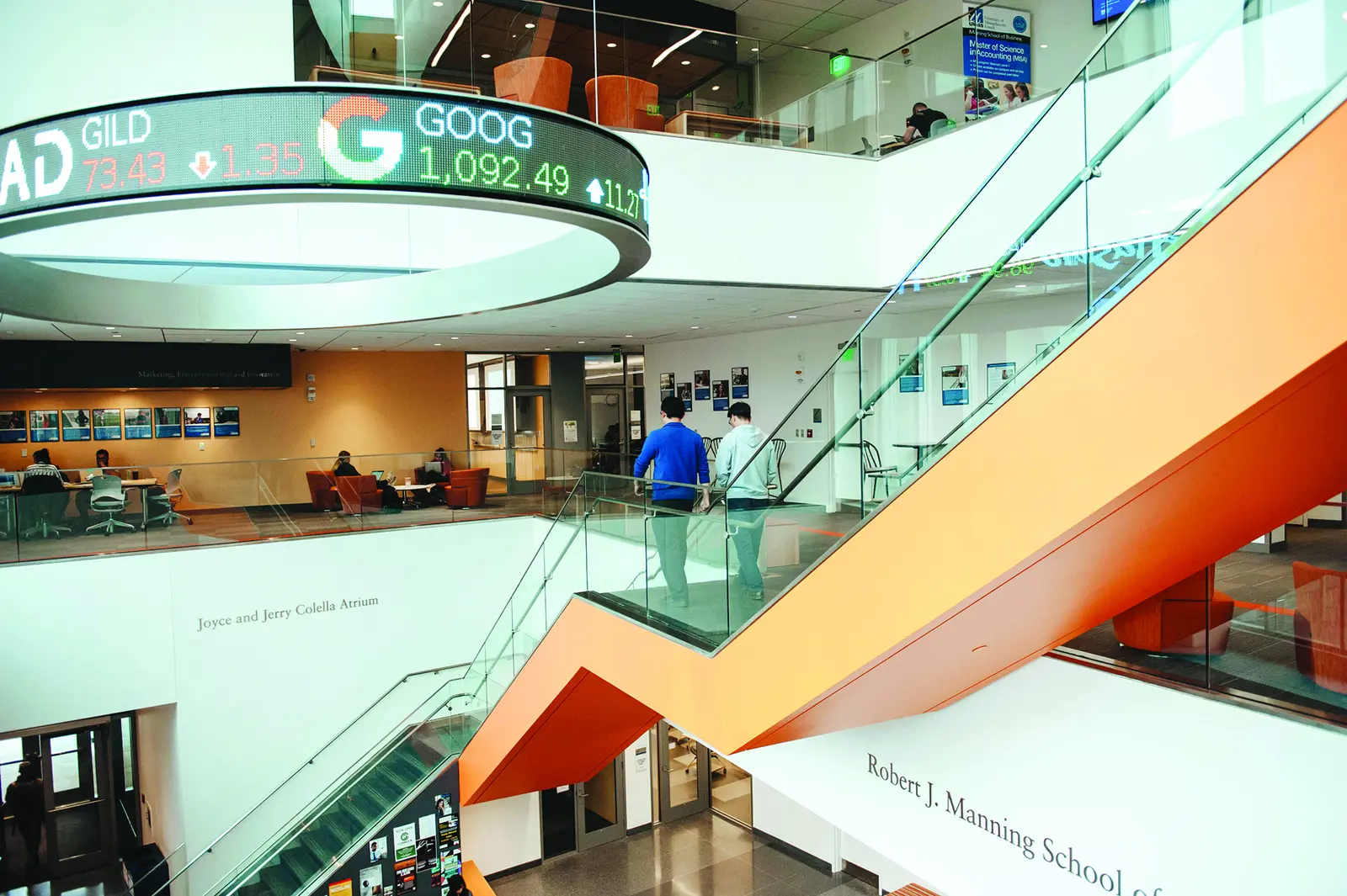 Two students walk down staircase in open atrium with stock exchange prices on round display over lobby