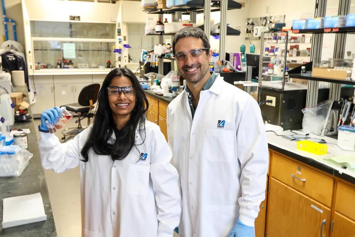 A young woman and a man wear lab coats and safety glasses while posing for a photo in a chemistry lab.