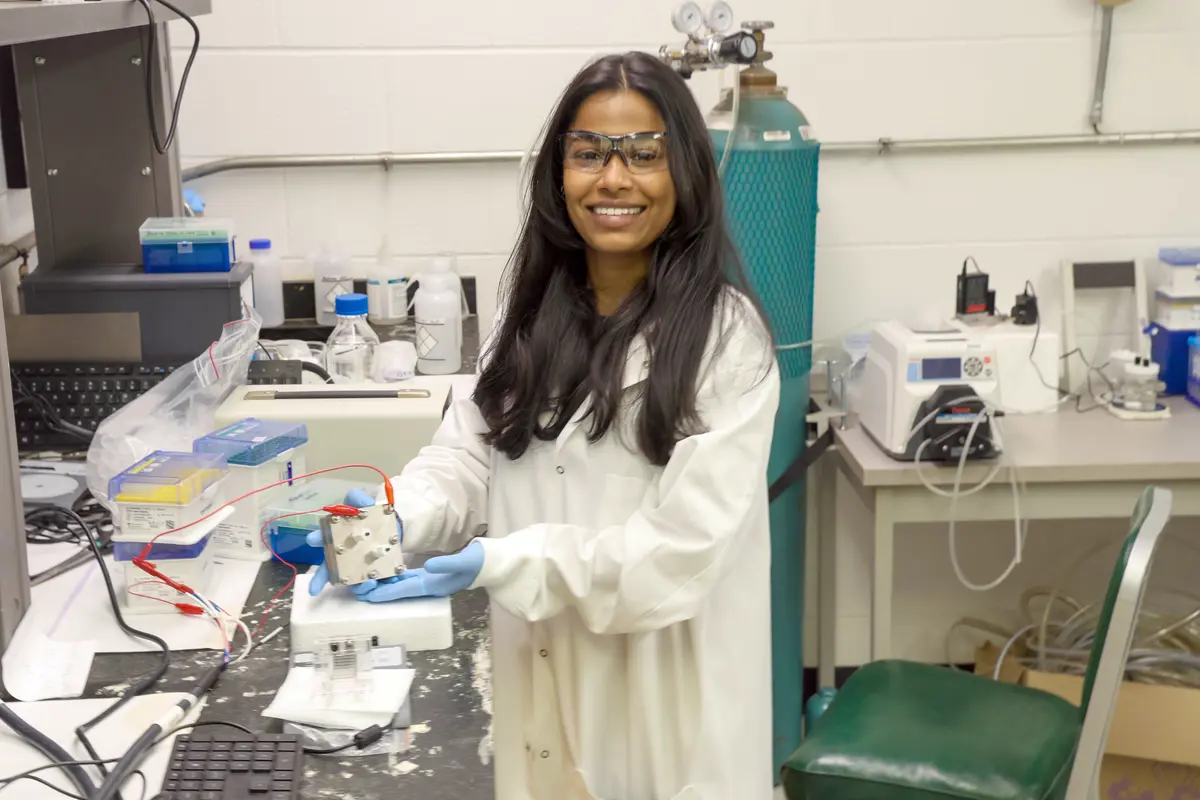 A young woman in a lab coat and safety glasses poses for a photo in a chemistry lab.