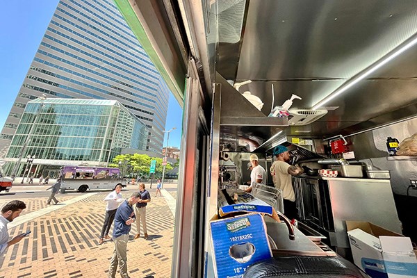 Three young men work in a food truck while customers wait outside in a plaza