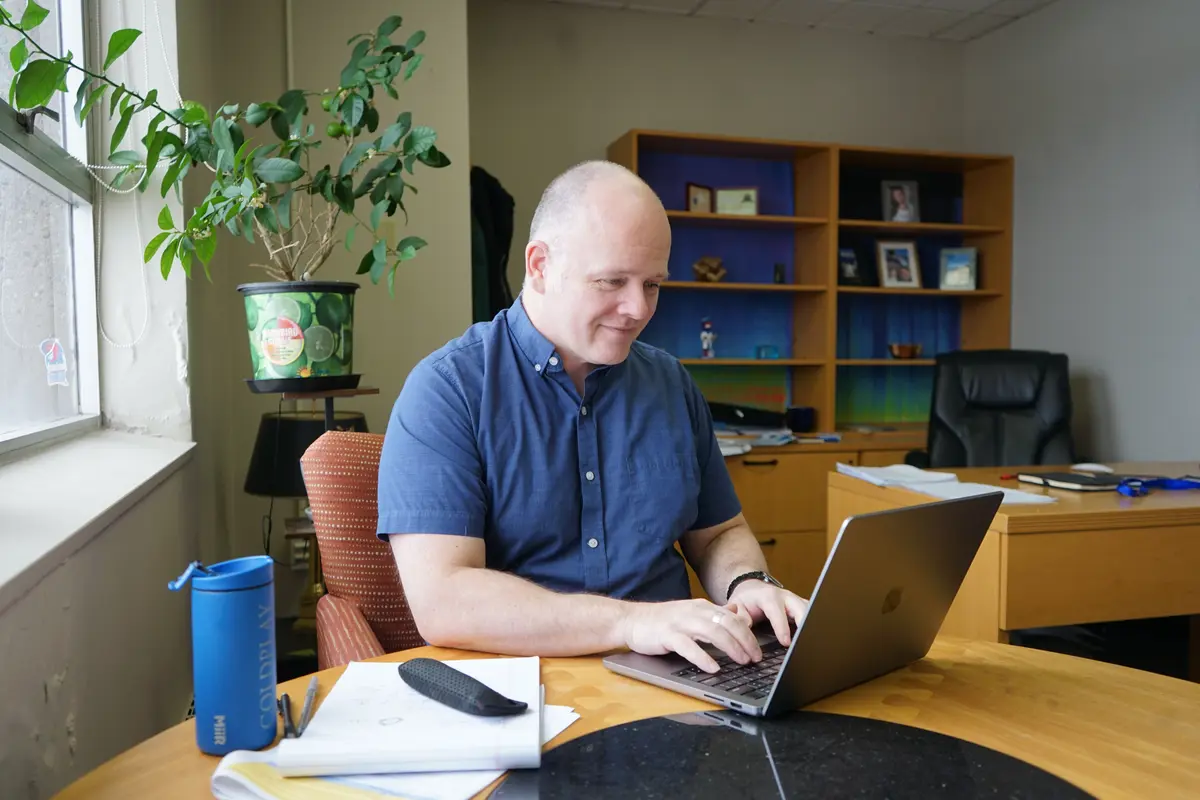 A man in a blue shirt works on a laptop while sitting at a table near a window in an office.