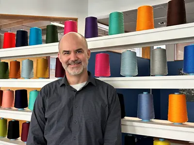Paul Kuttner stands in front of large spools of colorful cotton thread for weaving