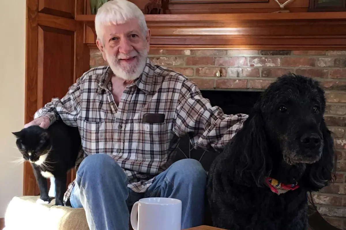 A man with white hair and a white beard poses for a photo with a cat and a dog in front of a fireplace.