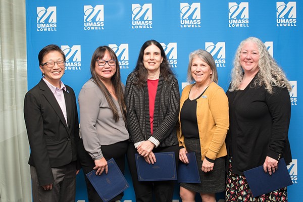 Five women pose for a photo in front of a UML backdrop