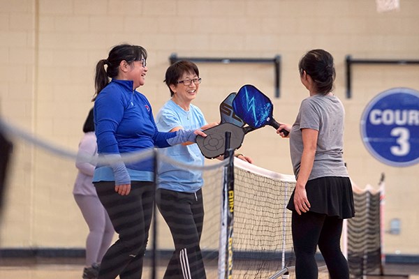 Three women tap pickleball paddles together at the net following a game