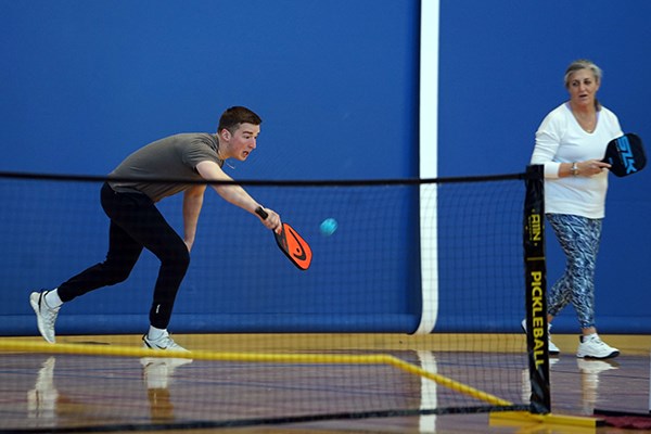 A young man lunges to hit a backhand in pickleball while his teammate looks on