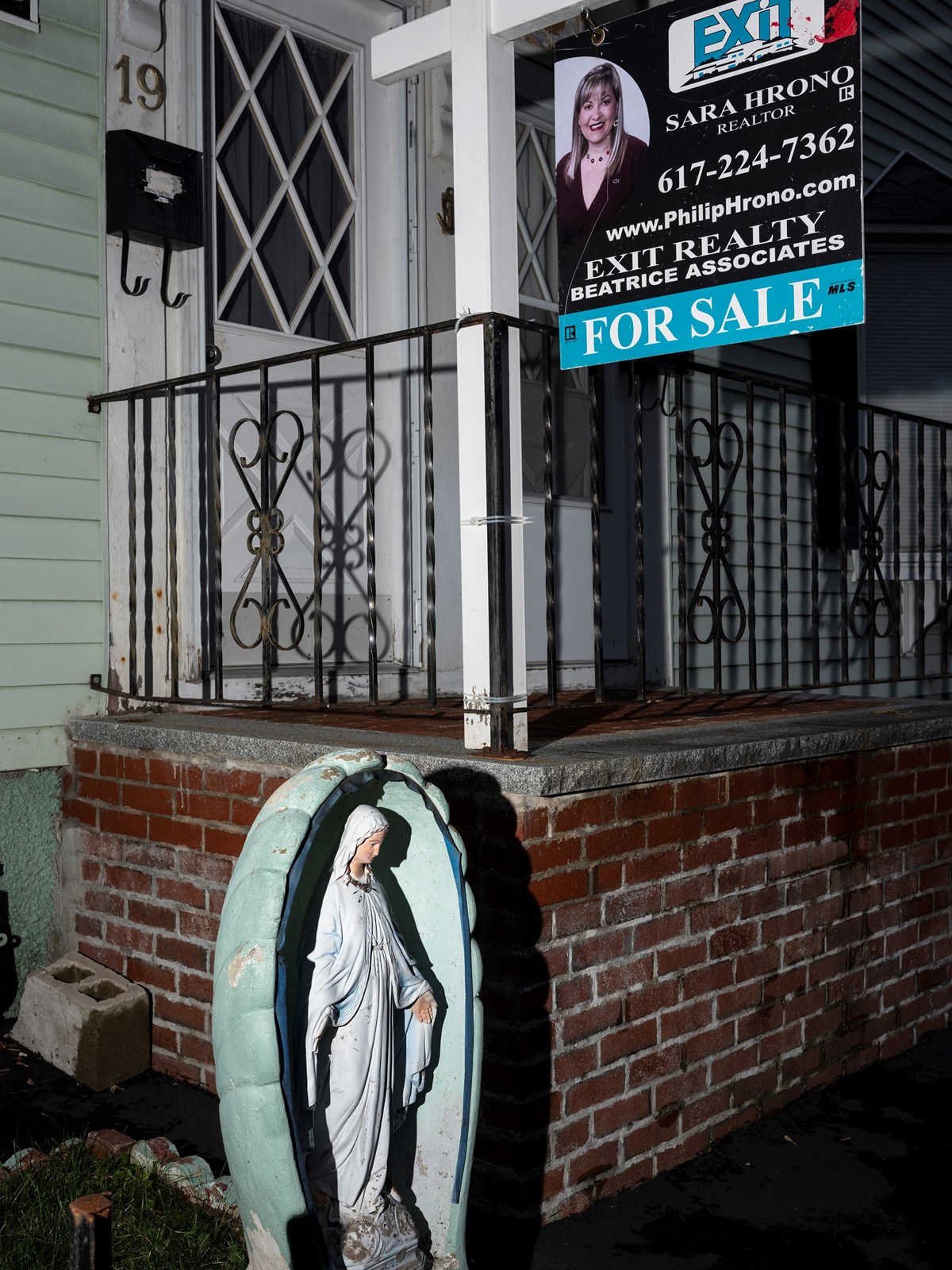 Picture of the outside / entrance to Lowell home with a for sale sign and the realtor's information as well as a statue of Mary.