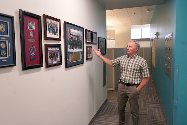 A man in a plaid shirt gestures toward a wall with framed police memorabilia