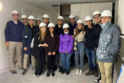 A dozen students wearing white hard hats pose for a photo in building under construction